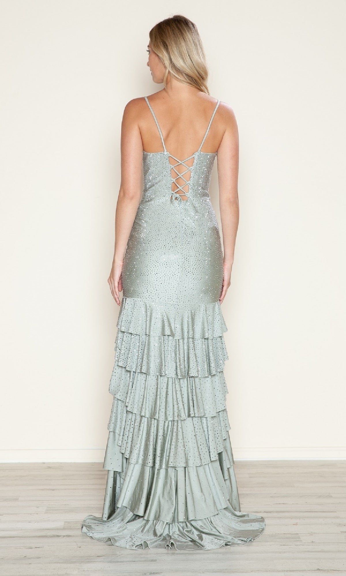 A woman stands on a wooden floor in the Poly USA 9620 Strappy Long Ruffled Prom Dress, featuring thin straps, a lace-up open back, shimmering sequins, and a layered ruffled floor-length skirt against a plain wall.